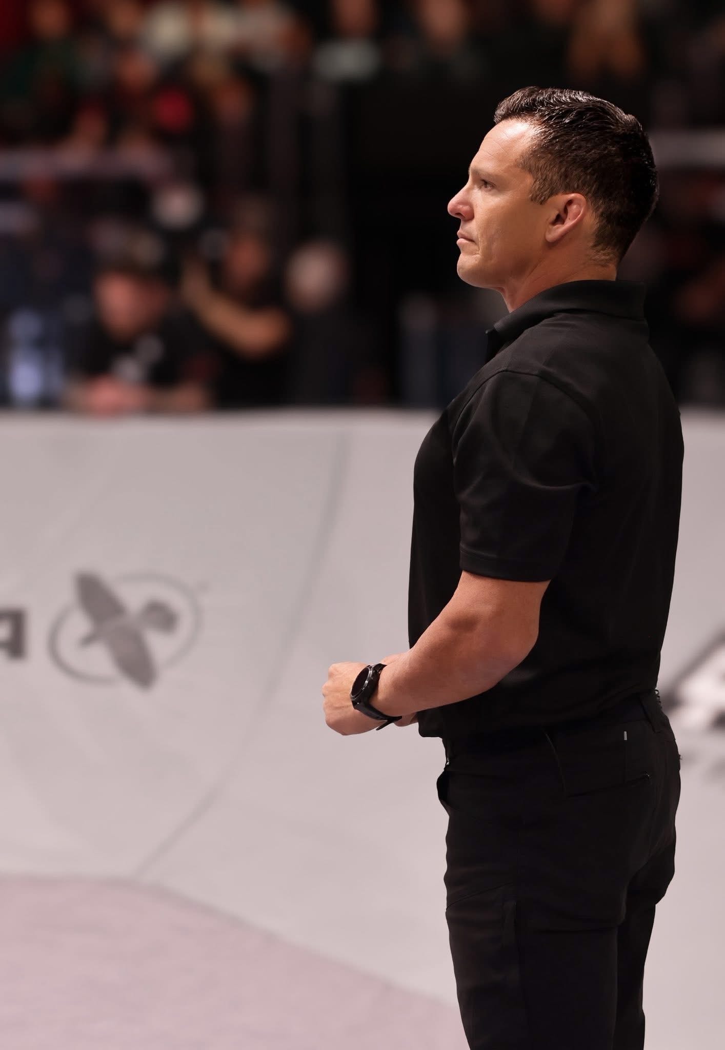 UFC Referee Jason Hertzog standing tall in profile, arena crowd blurred behind him